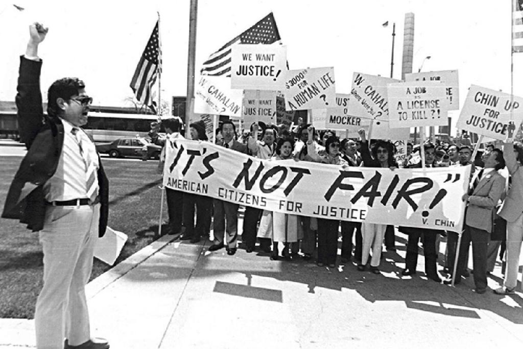 Demonstrators standing with signs protesting for justice for Vincent Chin. A large banner spans in front of them, reading "It's not fair!" in all caps, followed by "American Citizens for Justice." Courtesy of the collections of Helen Zia and the Vincent and Lily Chin Estate. 
